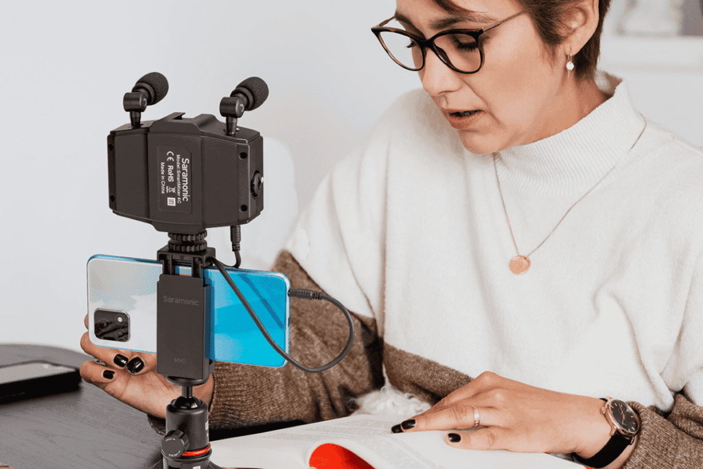 Mulher fazendo a leitura de seu livro enquanto grava com o celular, com microfones acoplados.