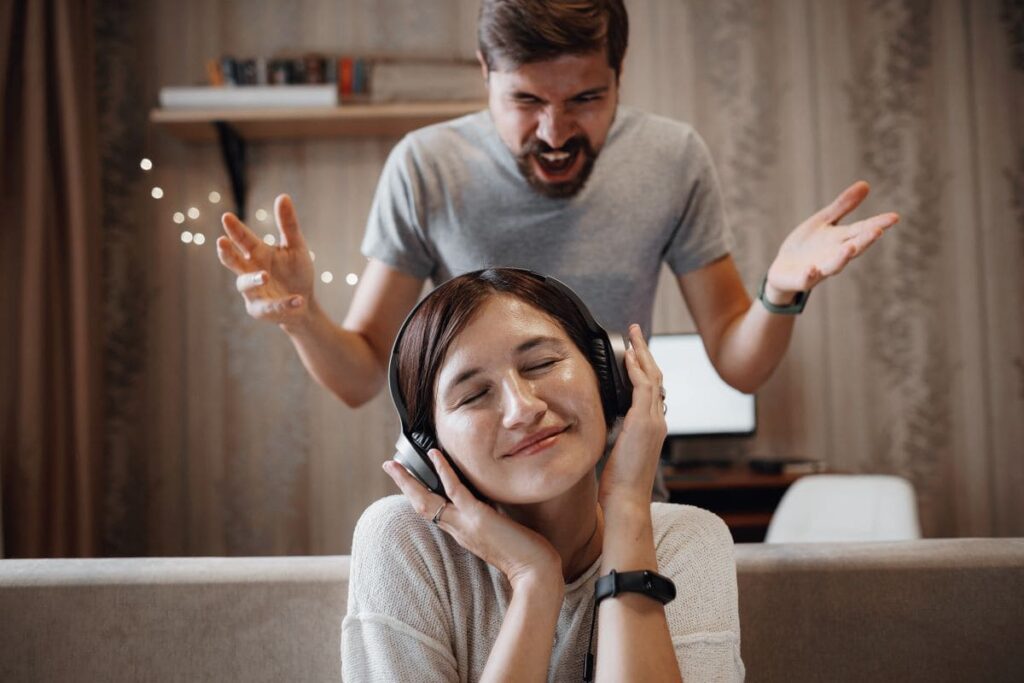 homem zangado gritando com a mulher sentada no sofá e cobrindo as orelhas com fones de ouvido.