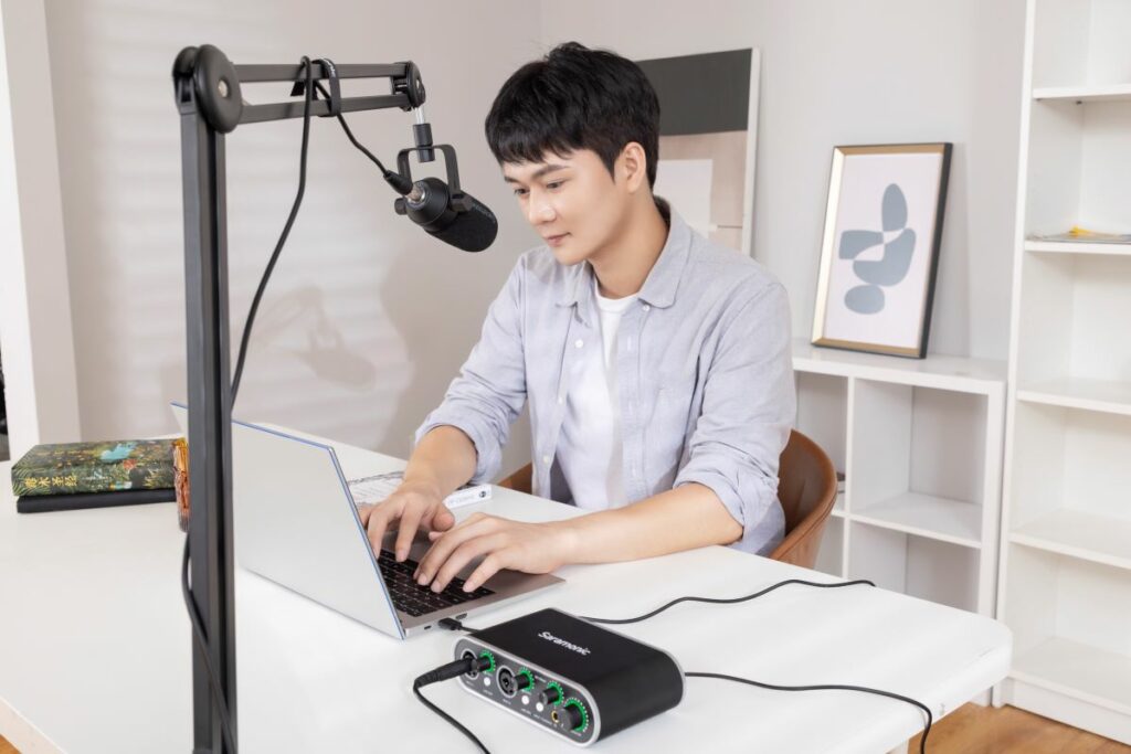 Homem sentado à sua mesa digitando em seu notebook, com o MV-Mixer Saramonic ao lado e um microfone acima de sua cabeça.