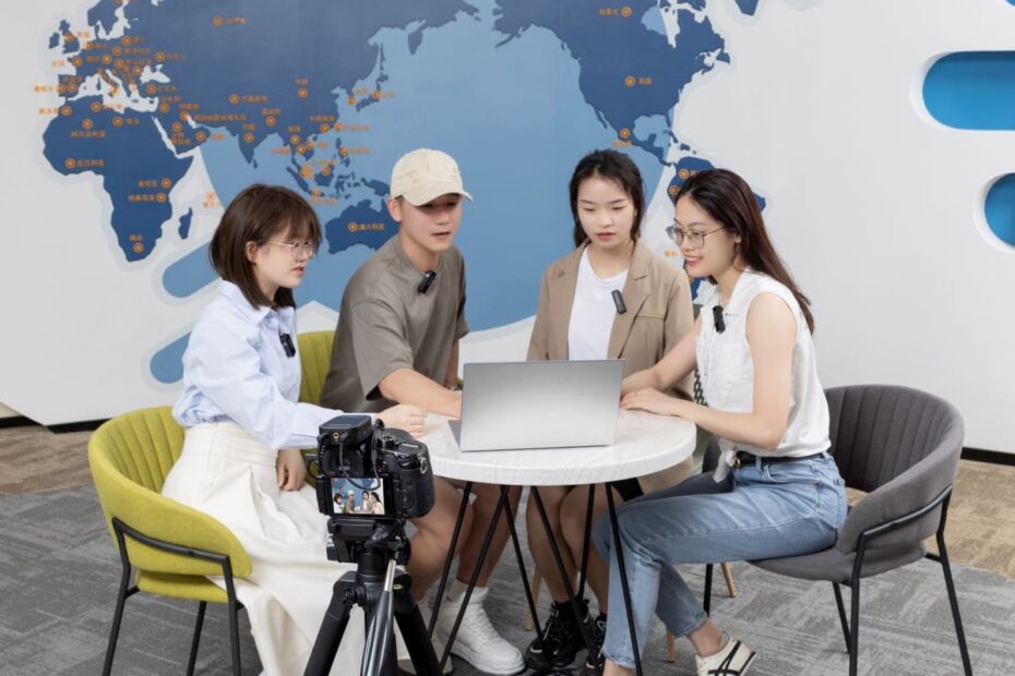 A imagem mostra um grupo de quatro pessoas reunidas em torno de um laptop, usando microfones de lapela sem fio, possivelmente para uma gravação de vídeo ou transmissão ao vivo. Há uma câmera em primeiro plano gravando a cena, sugerindo que estão criando conteúdo multimídia. No fundo, há um mapa do mundo, indicando um ambiente de trabalho colaborativo ou educacional com um alcance global.