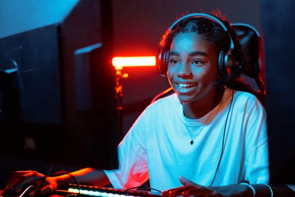 A imagem mostra um adolescente sorrindo enquanto joga videogame. Ele olha para a tela do computador e conversa online com seu clube de videogame. O ambiente possui uma iluminação azul e vermelha, e o teclado também é iluminado.