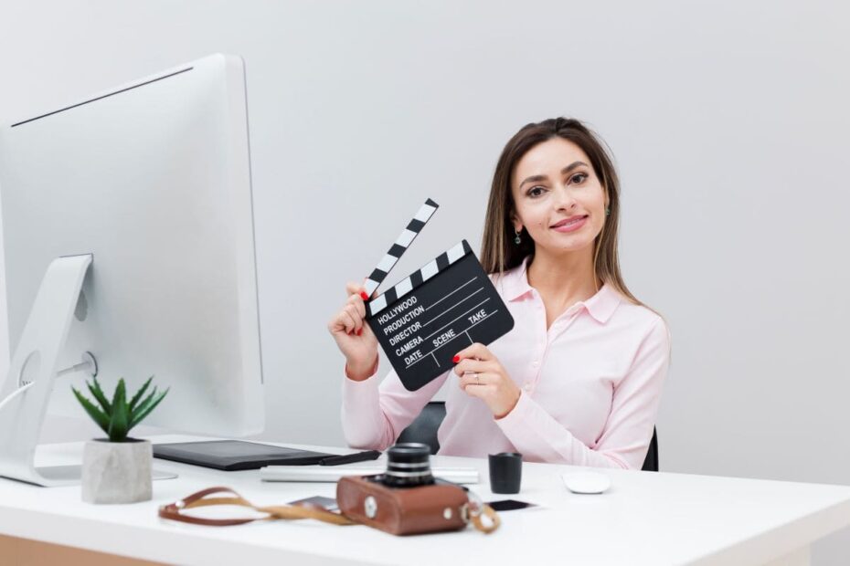 Na imagem, vemos uma vista frontal de uma mulher sentada à mesa, segurando uma claquete, pronta para iniciar uma gravação. Ela está focada e preparada para utilizar o audiovisual como ferramenta de vendas.