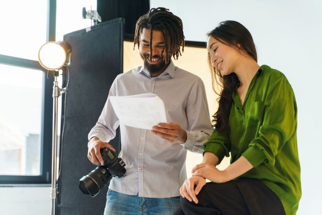 A imagem retrata dois profissionais em um estúdio de fotografia, engajados em uma discussão colaborativa. O homem segura uma câmera e um papel, sugerindo que estão revisando ou planejando aspectos de um projeto audiovisual. Esta imagem é ideal para ilustrar a importância do briefing no processo criativo e produtivo de projetos audiovisuais.