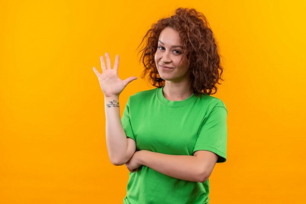 Imagem de uma jovem mulher com cabelo curto e cacheado, vestindo uma camiseta verde, sorrindo e mostrando a mão levantada com cinco dedos abertos, em pé contra uma parede laranja.