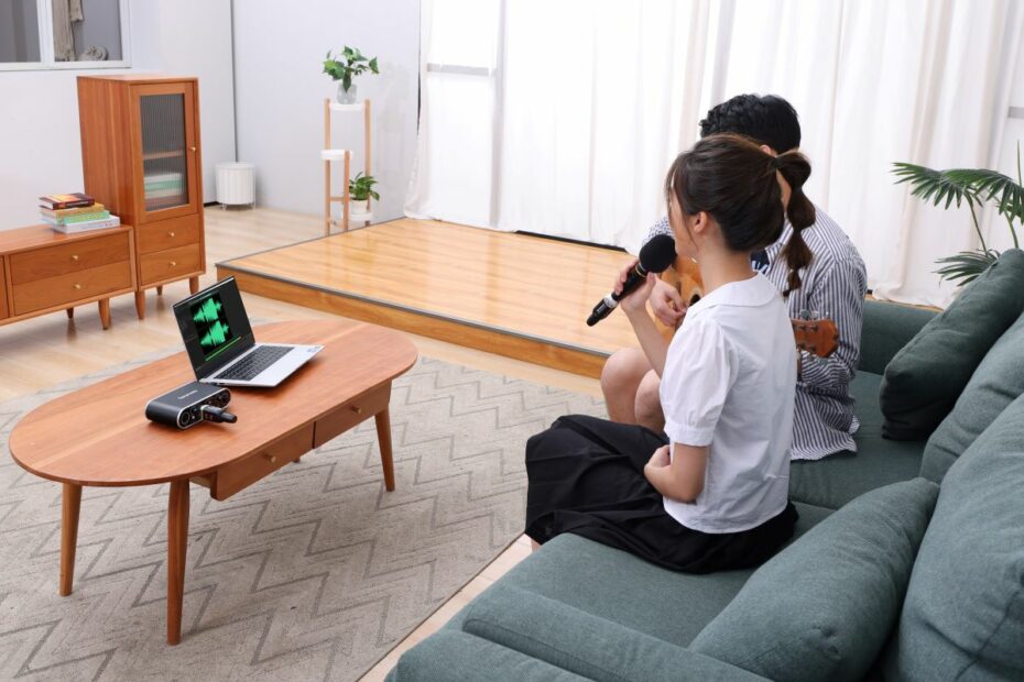 Um homem e uma mulher estão sentados no sofá em frente ao notebook, gravando com o interface de áudio MV-Mixer da SARAMONIC. O homem está tocando violão enquanto a mulher segura o microfone próximo à boca.