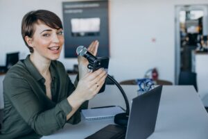 Mulher com cabelo curto sorrindo, segurando um smartphone e usando um microfone em uma mesa com um laptop e um notebook.