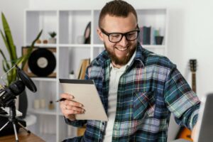 Homem sorridente com tiro médio olhando para tela do computador e segurando um caderno em frente a um microfone de mesa.