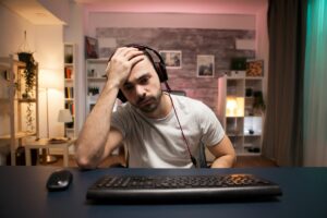 Um homem usando fones de ouvido está sentado em uma mesa com um teclado e mouse, apoiando a cabeça na mão. A sala tem estantes de livros, plantas e iluminação suave, criando uma atmosfera aconchegante. Ele parece estar imerso em pensamentos ou frustrado.